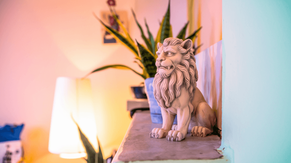 A close-up of a decorative lion statue on a ledge in a warmly lit apartment. The statue has intricate details and is accompanied by green indoor plants in the background, a soft-glow floor lamp, and cozy decor elements, creating a stylish and inviting ambiance.